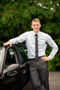 Jakim kierowcą jesteś, test, czyli młody mężczyzna w blond włosach, w szarych spodniach, jasnej koszuli i ciemnym krawacie, stoi przy ciemnym samochodzie i opiera się o niego prawą ręką