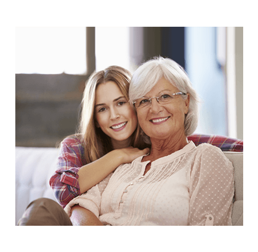 Babcia, czyli kobieta z siwymi włosami w okularach i jasno różowej bluzce z wnuczką w długich włosach, w koszuli w kratę, siedzące na kanapie i patrzące z uśmiechem przed siebie