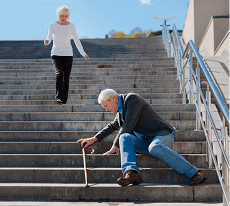 Starszy mężczyzna ubrany w jeansy i szary sweter próbujący z laską wstać ze schodów i biegnąca do niego po schodach kobieta ubrana w ciemne spodnie i białą bluzkę